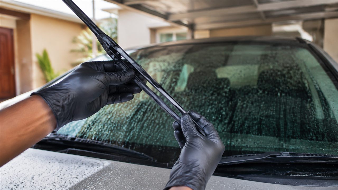 Gloved hands inspecting a windscreen-wiper blade above a rain-speckled windscreen